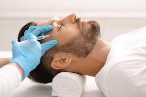 Cosmetologist making lifting procedures for handsome middle aged man at aesthetic clinic, side view. Man getting beauty injection against eye wrinkles from unrecognizable plastic surgeon photo