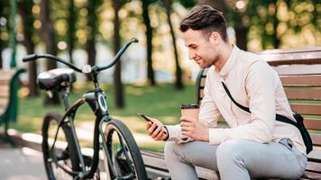 Work outdoors, take a break and get to office in morning. Smiling millennial guy manager sits on bench with bicycle, holds takeaway paper cup of coffee and reads message in gadget, in city, outdoor photo