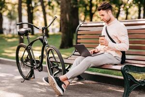 Work with pleasure. Side view of handsome man with stubble in casual clothes with bag, typing on laptop and drinking coffee takeaway, sitting on bench near bicycle in city park, outdoors, free space photo