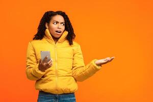 Displeased Black Woman Holding Smartphone, Having Problem With Cellphone Gesturing Aside At Copy Space Standing On Yellow Studio Background, Wearing Winter Clothes. Cellphone Issue, Bad News Message photo