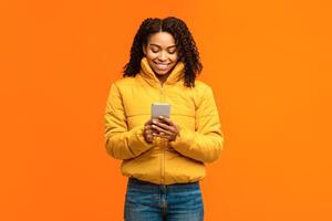 African American Woman With Smartphone Texting Standing Over Yellow Studio Background, Wearing Warm Winter Clothes. Mobile Phone User Using Cellphone Application Concept photo