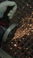 Construction worker cutting metal surface with angle grinder creating bright flying sparks, close-up view of gloved hands operating power tool in industrial workshop environment. Vertical video
