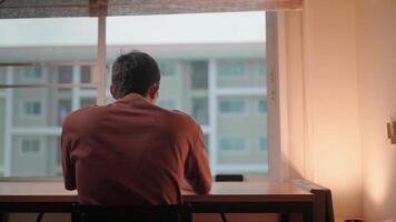 A solitary figure sits at a desk, back to the camera, gazing out a window. The soft light of the setting sun casts a warm glow on the scene. video