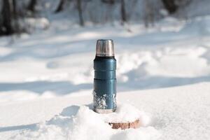 Metal blue thermos standing on a snowy tree stump in a winter forest on a sunny day photo