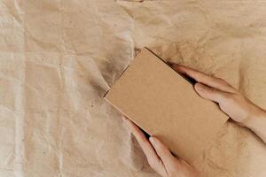 Hands holding brown cardboard box, wrapping paper background. Top view, layout. Place for logo, announcement, text. Gift, delivery concept photo