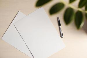 Blank sheet of paper and pen on beige table. Flatlay mockup. Part of a house plant in the frame. Place for text photo