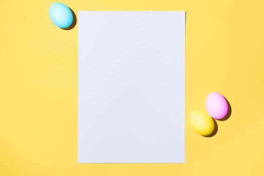 Easter layout. White blank sheet of paper and Easter colorful eggs on a yellow background, flat lay. Greeting card with place for text photo