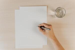 Blank paper, glass of water and hand holding pen, top view. Flatlay, mock up. photo