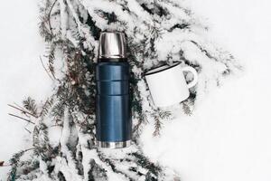 Thermos with a mug lying on a pine branch covered with snow. Top view. photo