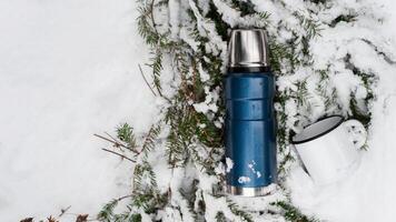Camping in the winter forest concept. Thermos and iron mug lying on a pine branch in the snow. Top view, copy space. Vacuum flask for travel. photo