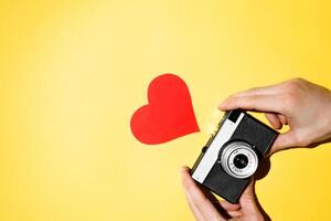 Hands holding vintage film camera, top view. Retro photo camera and red heart like on a yellow background, flat lay.