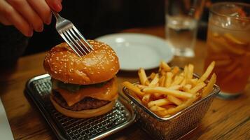 Close-up of a sesame seed burger served with cheese and french fries on a wooden table. A refreshing cold drink and a plate are also in view, creating a cozy dining experience. photo