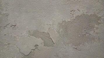 A close-up of a rough, worn-out concrete wall with a gritty texture, featuring cracks and crevices. The color is a dull, muted gray with subtle variations in shade, showing off the uneven surface. photo