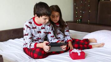 brother and sister, sit on the bed in the morning in pajamas and look at a digital tablet video