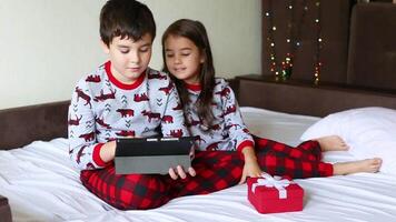 Beautiful brother and sister, sit on the bed in the morning in red pajamas and look at a digital tablet video