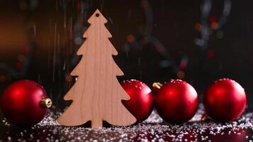 Snow falls on a wooden Christmas tree toy in the form of a fir on a dark table with red balls. video