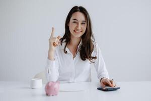 Woman in a white shirt smiling while pointing up at a desk with a piggy bank and calculator in a bright, minimalistic setting photo