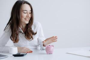 Young woman enjoying savings while interacting with a pink piggy bank on a desk in a bright office setting photo
