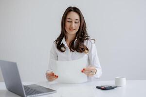 Woman in a white shirt reviews documents at a desk with a laptop and calculator in a bright office setting photo