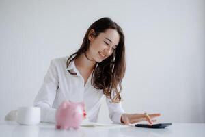 Young woman calculating finances at a minimalist desk with a piggy bank and notebook in a bright, modern office setting photo