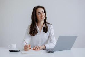 Young woman working at a desk with a laptop and notebook while smiling in a modern office setting photo