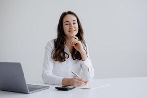 Professional woman smiling while taking notes at a desk with a laptop and calculator in a bright office environment during daytime photo