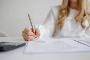 Individual completing paperwork with pen while sitting at desk with calculator and documents in bright workspace photo