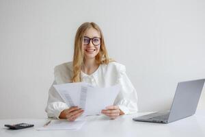 Woman reviewing documents at a desk with a laptop in a bright office environment during the day while smiling photo