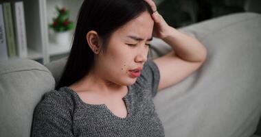 Handheld high angle view shot, Young Asian woman sit on the sofa alone, lost in thought. Her brows were slightly furrowed and expression tense in the quiet atmosphere at home video