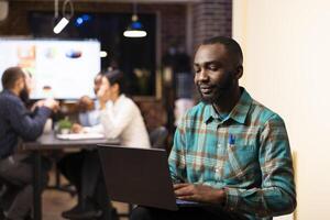 African american entrepreneur reviews financial planning data on device as startup team collaborates in background. Portrait of businessman with laptop, checking emails and planning his schedule. photo