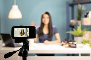 Selective focus on a camera mounted on a tripod, recording a beauty vlogger. The device screen shows a smiling asian woman waving as she begins her live broadcast on makeup tutorials and reviews. photo