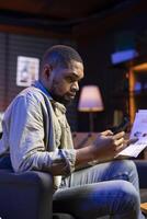 Black remote worker doing math calculations using financial graphs on papers, working from his cozy loft apartment. Self employed person comparing business economic figures on files. photo