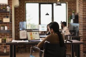 Female employee in a wheelchair working on a laptop, reviewing financial reports and data charts in a startup office. Asian businesswoman using her personal computer in a modern, inclusive workspace. photo