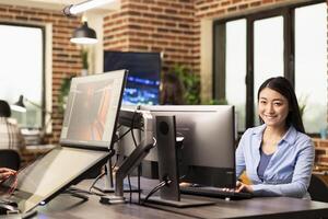 Portrait of asian businesswoman sitting at table and happily working on her computer in modern workspace. Cheerful female professional with her hands on keyboard, typing and looking at camera. photo