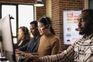 Multiethnic call center agents sit at their workstations, handling business inquiries and communicating with remote customers. Coffee, computers, and headsets set the office scene. photo