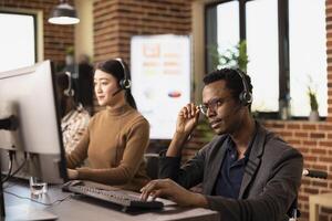 Male wheelchair user touching his glasses and working attentively at his office desk. Black businessman using a headset to communicate with online clients and entering their details on computer. photo