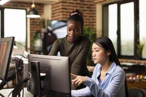 Focused asian and african american coworkers looking at computer, analyzing and discussing on updating graphic designs. Diverse female editors in conversation about digital art in modern office. photo