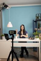 Beauty influencer sits at desk in a studio with a blue backdrop, engaging with her online audience during livestream. Perfect for themes of vlogging, content creation, and influencer marketing. photo