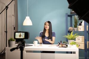 Asian woman filming a livestream makeup tutorial in a studio setup. Beauty vlogger talks to her online audience with a camera on tripod and bright lighting, reviewing cosmetics at her desk. photo