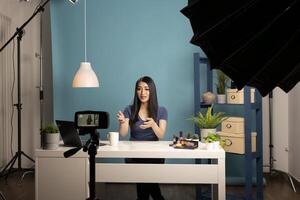 Young asian blogger recording a cosmetic review in a studio with a camera on a tripod. She sits at a desk with makeup products, using professional lighting and a blue background. photo