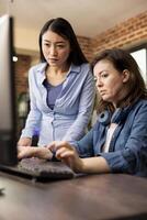 Two diverse women in modern office engaged in a creative discussion, one typing on the keyboard as the other reviews the work on the screen. Asian and caucasian colleagues collaborating on a project. photo