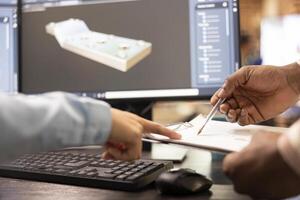 Closeup of multiethnic developers reviewing a sketch plan with 3D software displayed on computer monitor. Selective focus on diverse hands touching a clipboard, analyzing tool sizing numbers. photo