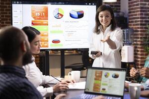 Smiling businesswoman reviewing financial charts with team in startup office. Coworkers are engaged in collaborative efforts, using technology to analyze future projects and discuss marketing strategy photo