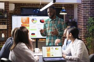 African american manager discussing with coworkers during late night meeting in brick wall workspace. Multiethnic colleagues using digital devices in reviewing marketing strategy in startup office. photo