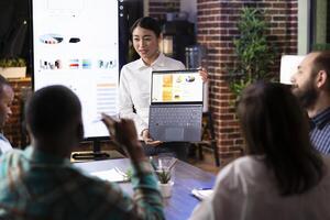 Asian financial advisor confidently leads a workplace discussion, presenting financial data on laptop. The modern office setting, digital screen and engaged team reflects dynamic business environment. photo