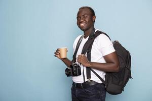 Excited male photographer stands against blue background, holding coffee cup with camera around his neck, ready for a client photoshoot. Black man with his photography gear looks optimistic in studio. photo