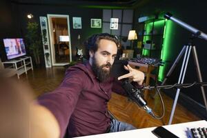 Influencer in home studio using smartphone on selfie stick to record live broadcast for online streaming platforms, talking with fans. Smiling media star filing himself doing cool hand gesturing photo