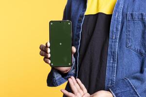 Confident casual person pointing at the copy space screen on gadget, pointing at the display on the mobile device. Young indian guy gesturing against orange background in studio. photo