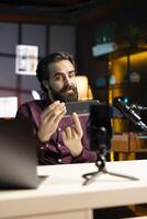 Tech guru informing audience about newly released mini soundbar. Man in studio pressing buttons on high end music playing device, testing specifications, filming with smartphone photo