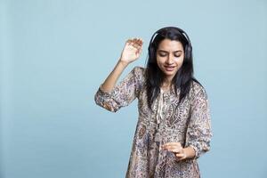 Relaxed happy woman playing music and having fun moving around, pretending to use turntables and dancing on the rhythm. Upbeat DJ wearing headphones, producing beats over studio background. photo
